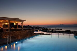 A poolside area with a stone wall, a covered seating area, and a view of the sea and distant mountains at dusk. - Summer Senses Luxury Resort (Paros)