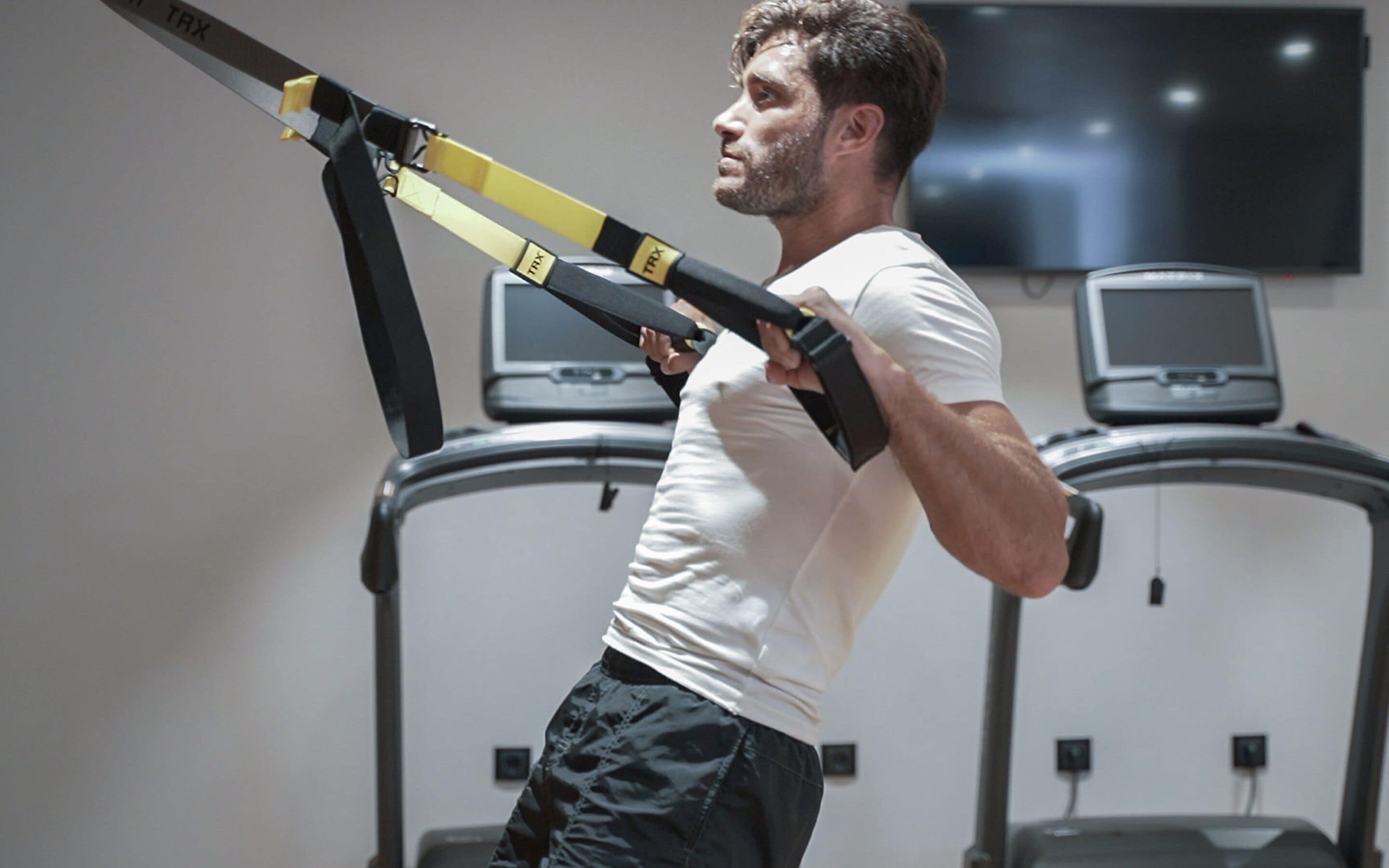 Um homem utilizando um treinador TRX em uma academia, com esteiras e uma televisão fixada na parede.