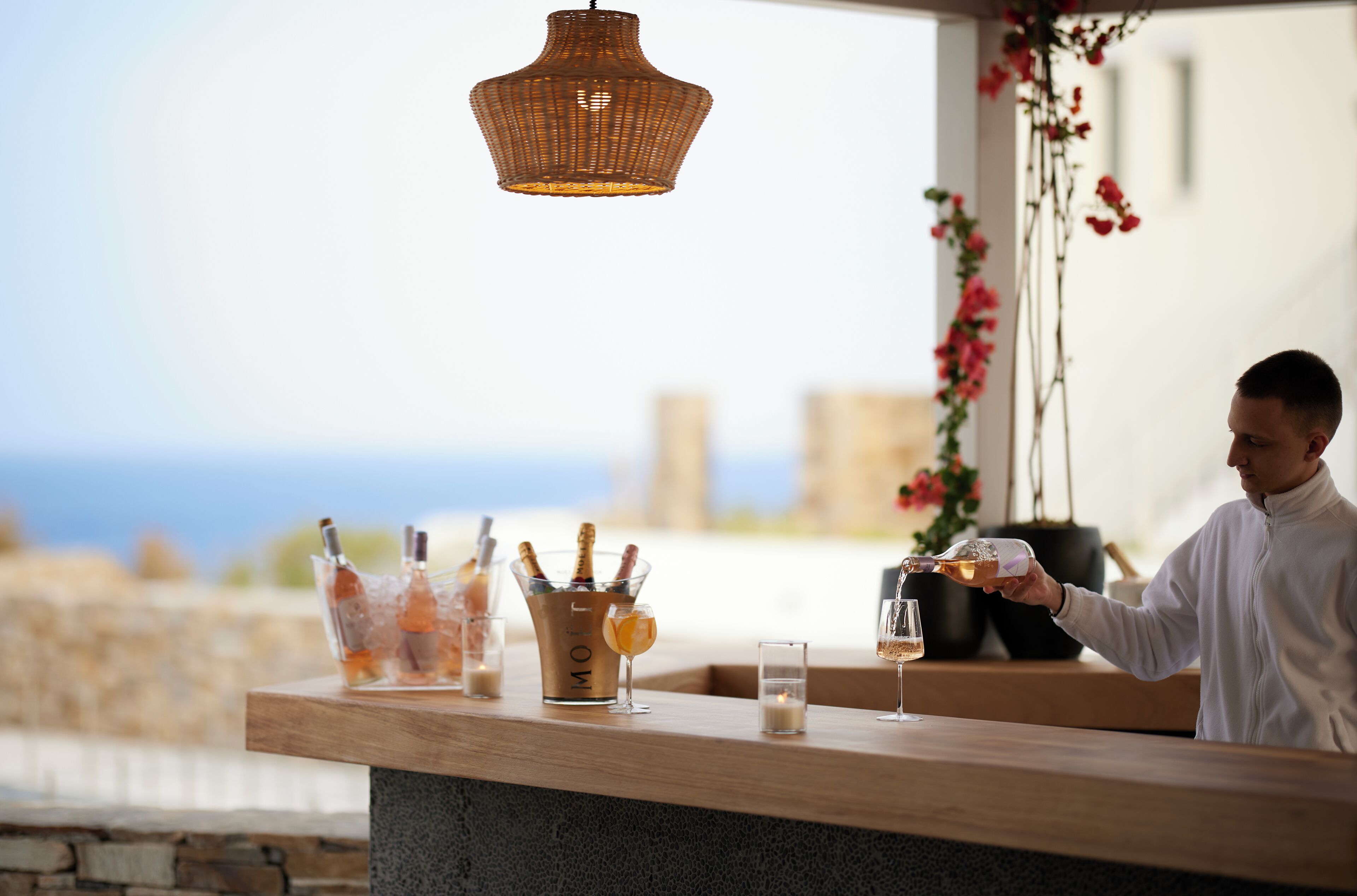 A bar area with a wooden counter, a wicker pendant light, and a view of the sea.