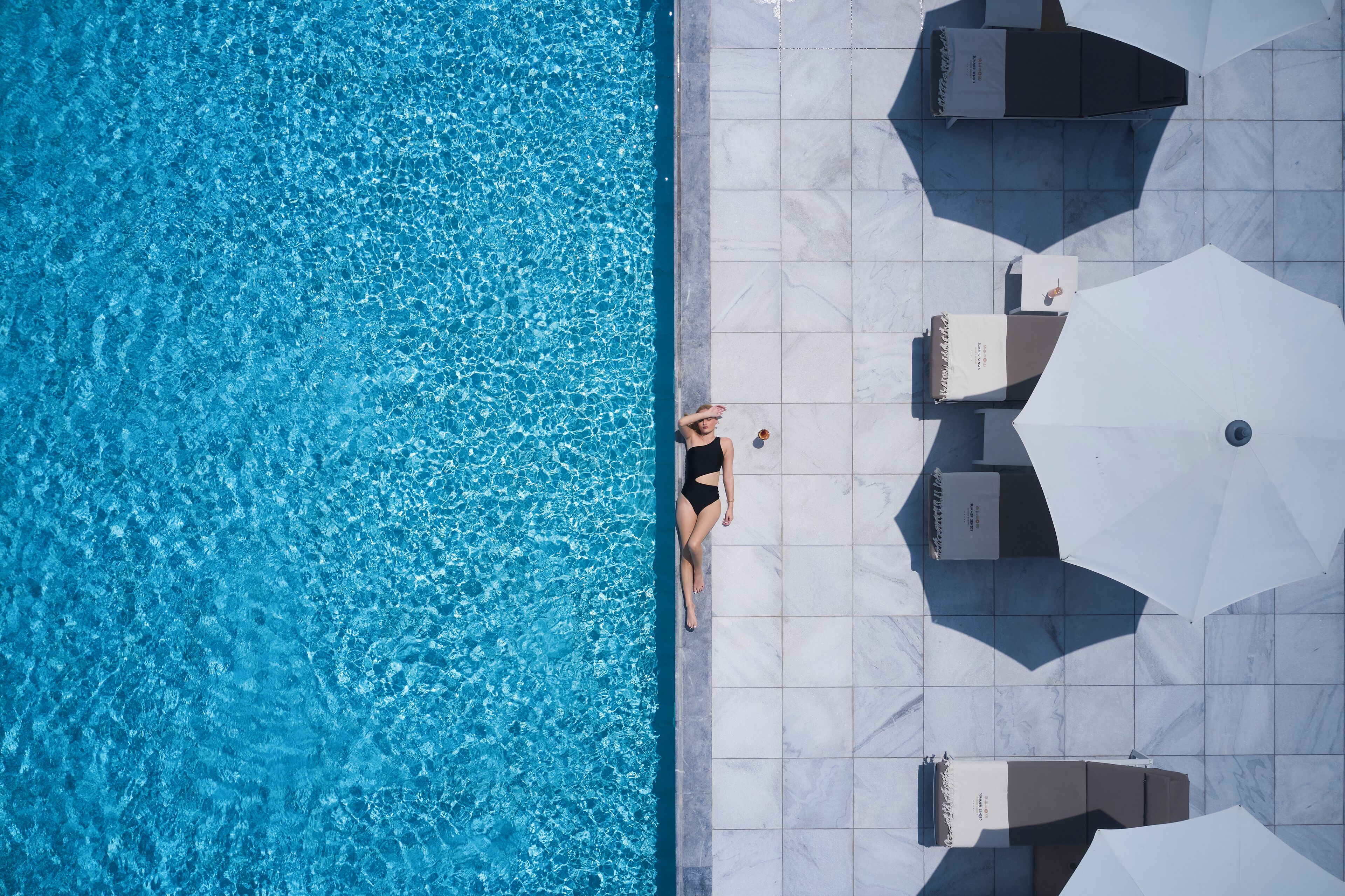 Vista aérea de uma piscina com uma pessoa de fato de banho, piso de azulejos e guarda-sóis.