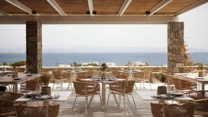 An outdoor dining area with stone pillars and wicker furniture, overlooking a scenic view of the sea and distant buildings.