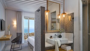 A modern hotel room with a large bed, a sitting area with a chair, a large window with a view, a bathroom with a large mirror, a sink, and a shower.
