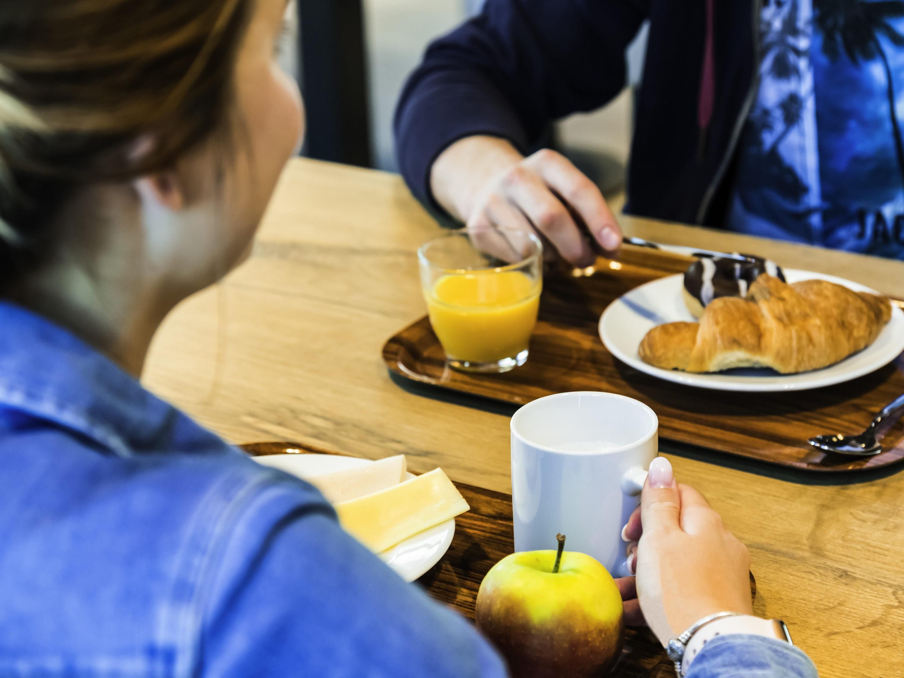 A dining table with a tray containing a glass of orange juice, a croissant, a plate with a croissant, a white mug, and an apple.
