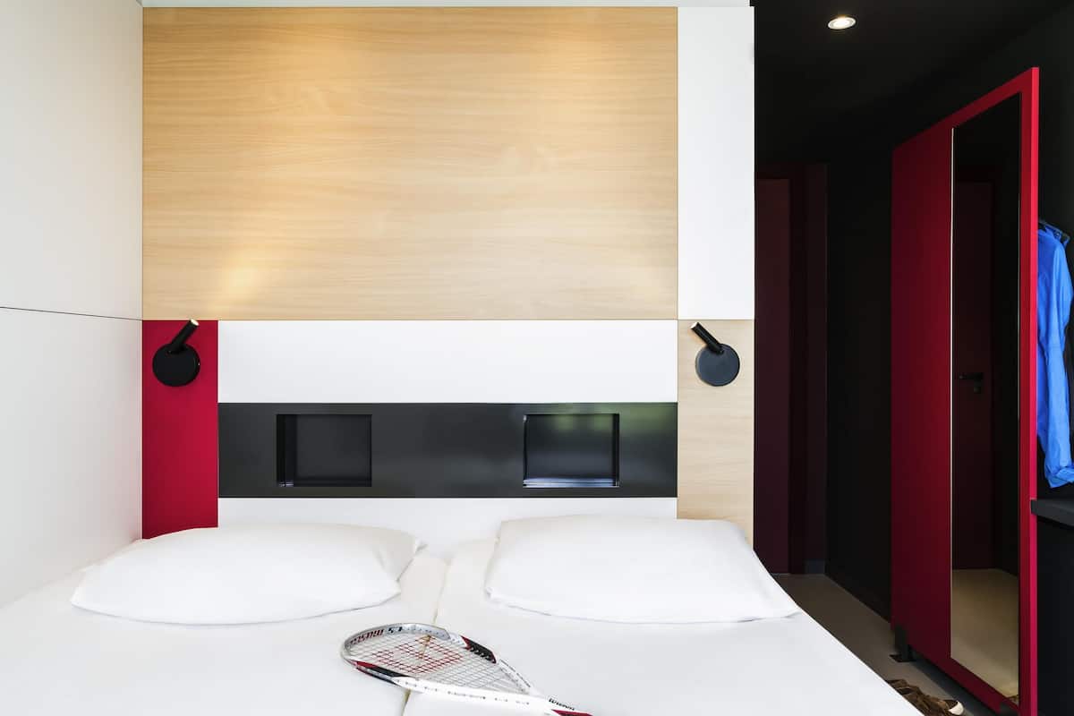 Una habitación de hotel moderna con cabecera de madera, cama con ropa blanca, una raqueta de tenis sobre la cama y una puerta roja.