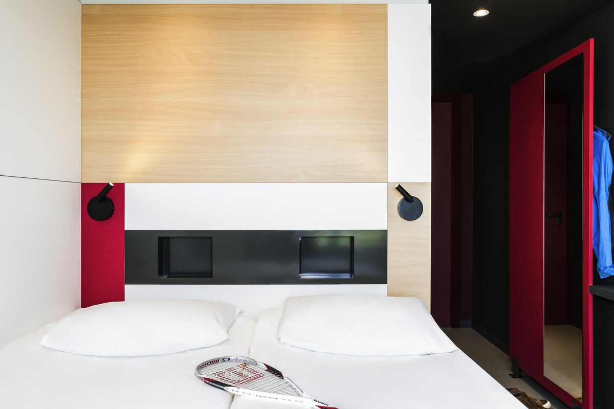 A modern hotel room with a wooden headboard, a bed with white bedding, a tennis racket on the bed, and a red door.