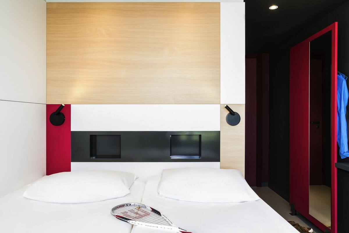 Une chambre d’hôtel moderne avec une tête de lit en bois, un lit recouvert de draps blancs, une raquette de tennis posée sur le lit et une porte rouge.