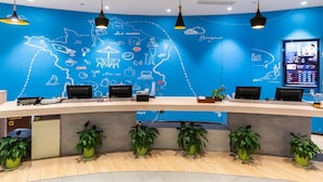 A reception desk with multiple computer monitors, a digital screen displaying information, and a blue wall with travel-related illustrations. - ibis Yixing South Renmin Rd (Wuxi)
