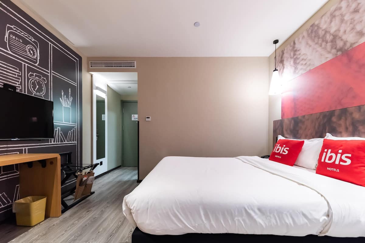 A hotel room with a bed, a TV, and a desk with a chair. There is a door leading to another room and a hallway.