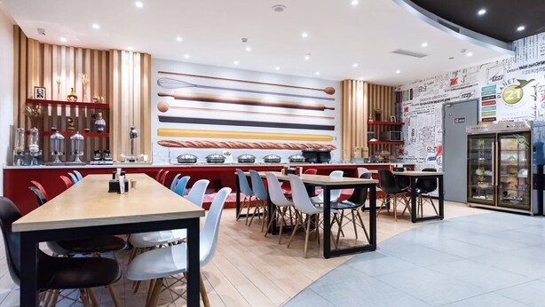 A modern restaurant with a long counter, multiple tables, and chairs. There are decorative items on shelves and a wall with various utensils displayed. - ibis Yixing South Renmin Rd (Wuxi)