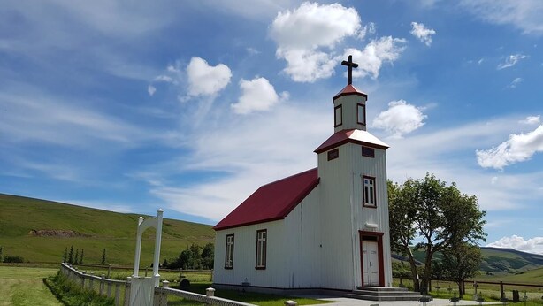Una iglesia blanca con techo rojo y una cruz en la parte superior, rodeada por una zona vallada y un campo cubierto de césped.