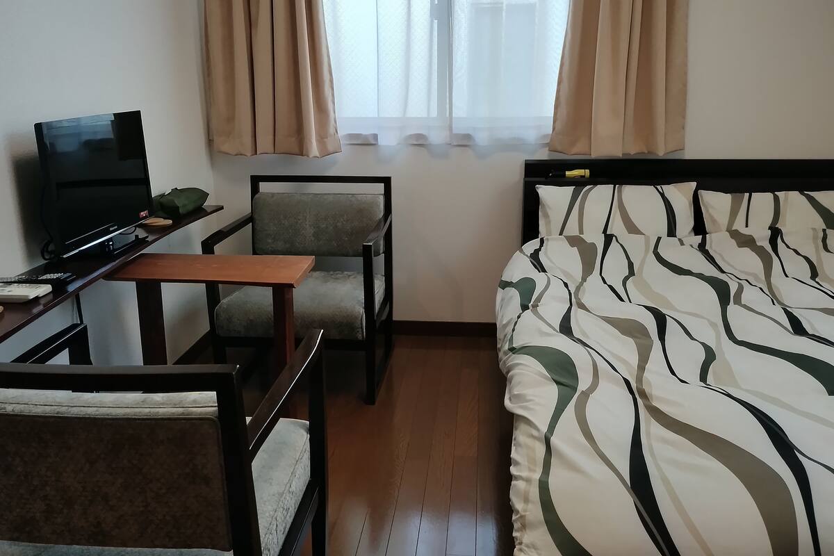 Habitació d'hotel amb un llit, una taula de treball amb una cadira, televisió i una finestra amb cortines.