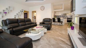 A modern living room with a sofa, armchair, and coffee table. There is a kitchen area in the background with a tiled backsplash and a flat-screen TV mounted on the wall. - Apartments Paradis (Rovinj)