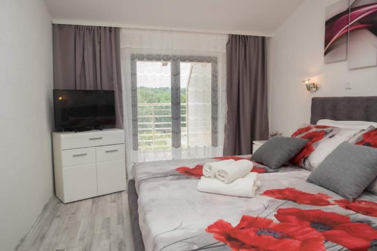 A bedroom with a bed, a TV on a stand, a window with curtains, and a view of greenery outside.
