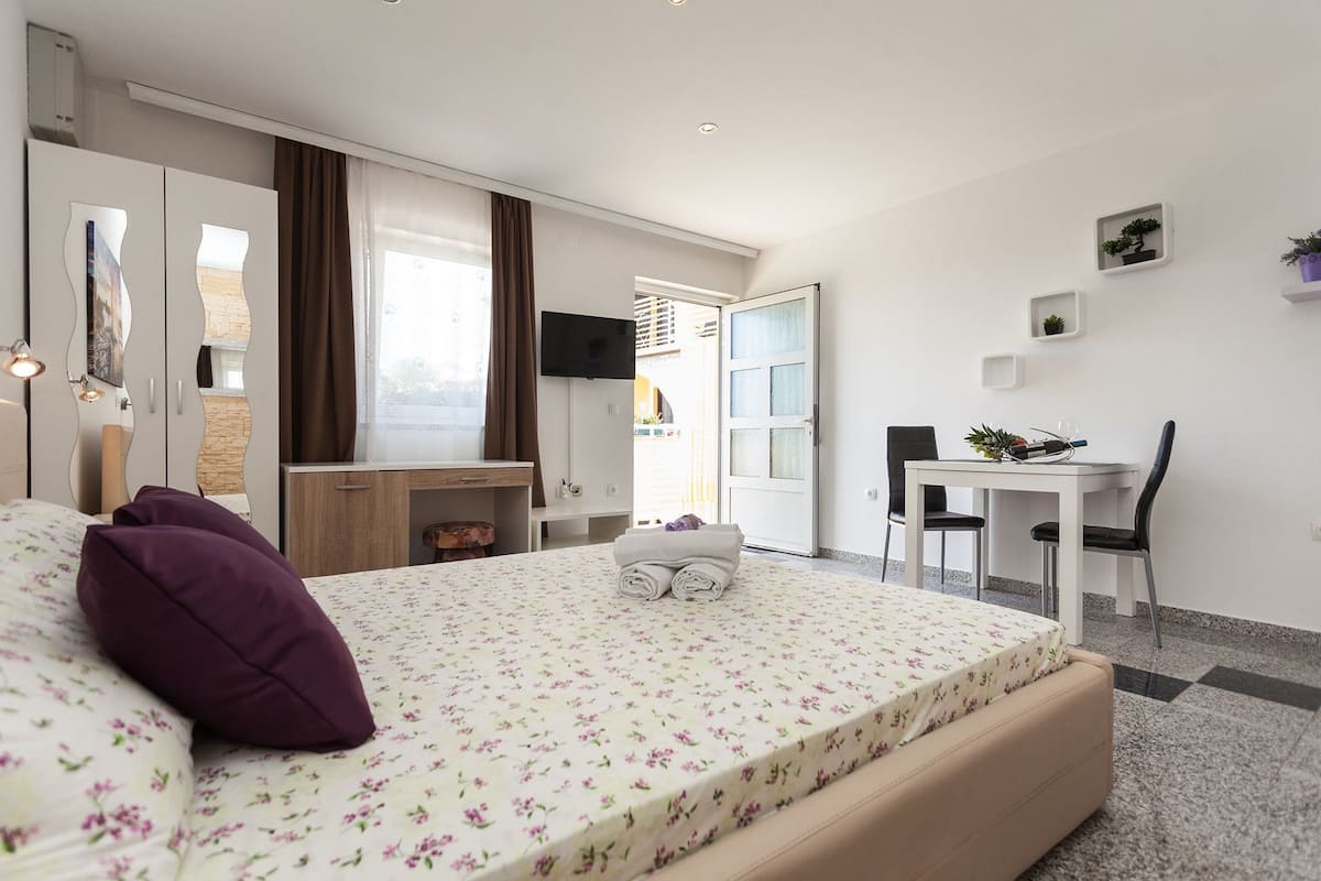 A bedroom with a bed, a desk, and a chair. There is a window with curtains, a flat-screen TV on the wall, and a small table with a plant.