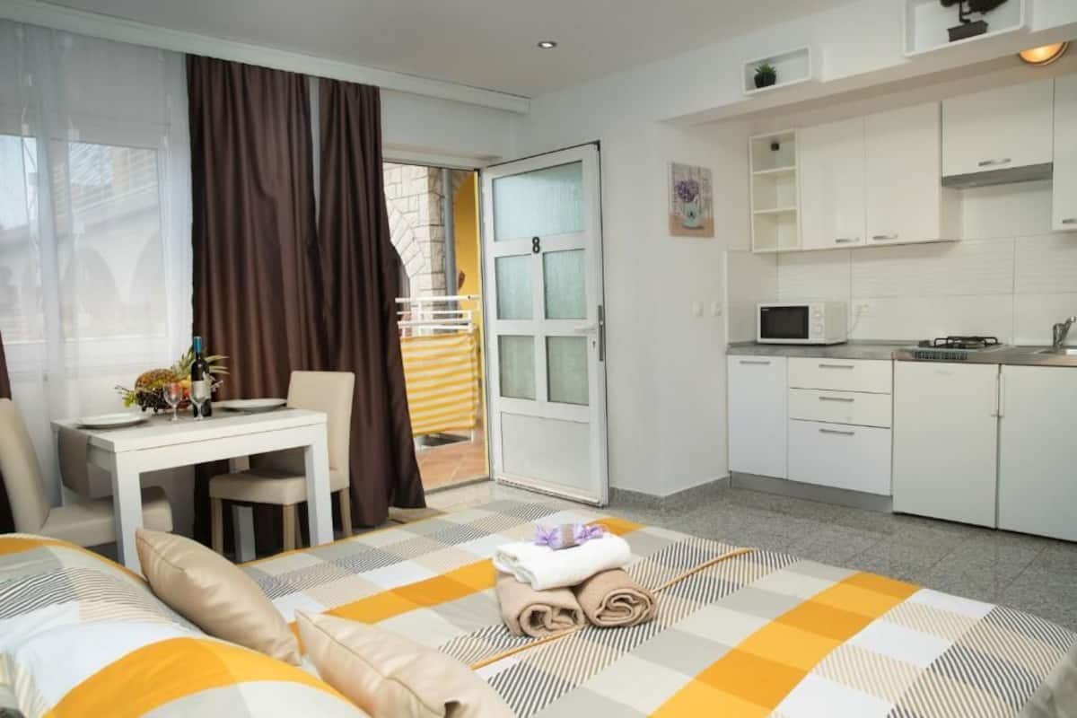 A hotel room with a bed, a desk, a chair, a kitchen area, and a bathroom.