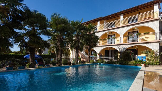 A swimming pool with a clear blue water surface, surrounded by palm trees and a building with balconies and arched windows. - Apartments Paradis (Rovinj)