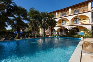 Une piscine à l’eau d’un bleu cristallin, entourée de palmiers et bordée d’un bâtiment doté de balcons et de fenêtres en arc de cercle.