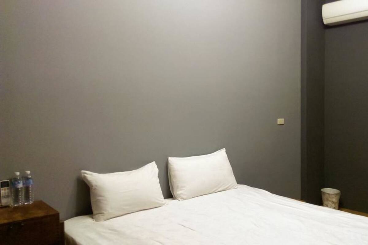 Habitación de hotel con cama, dos almohadas, mesita de noche con botellas de agua, un cesto de basura pequeño y un espejo en la pared.