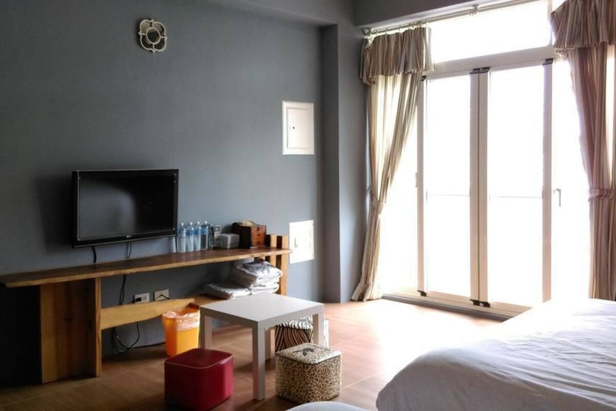 Habitación de hotel con cama, televisor sobre un soporte de madera, una mesita y una ventana con cortinas.