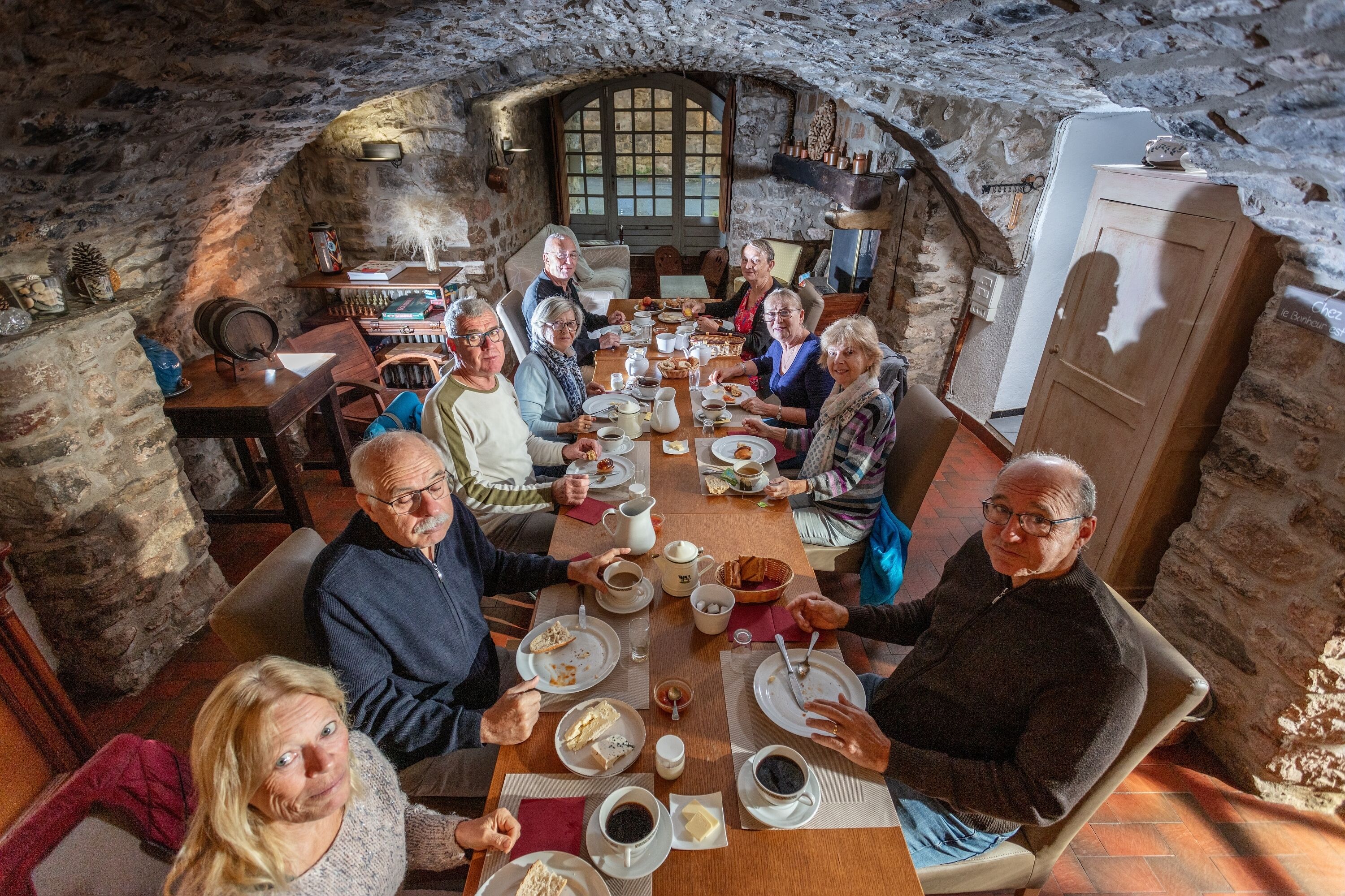 A stone-walled room with a high arched window, a wooden table with people dining, and a refrigerator in the corner.