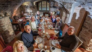 A stone-walled room with a high arched window, a wooden table with people dining, and a refrigerator in the corner.