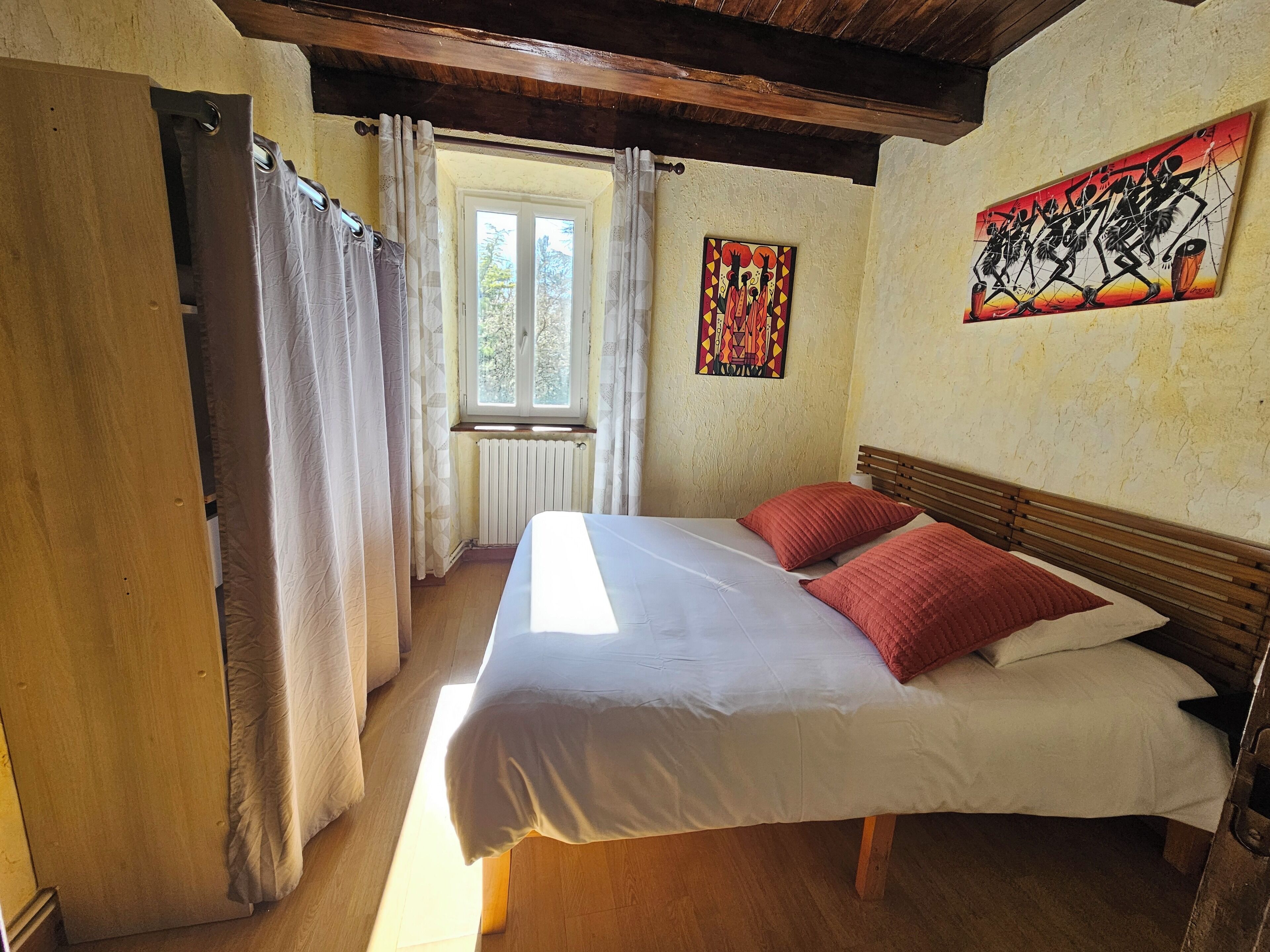 Ein Schlafzimmer mit einem hölzernen Bett, weißer Bettwäsche, roten Kissen, einem Fenster mit Vorhängen, einem Heizkörper und einem Wandbild.