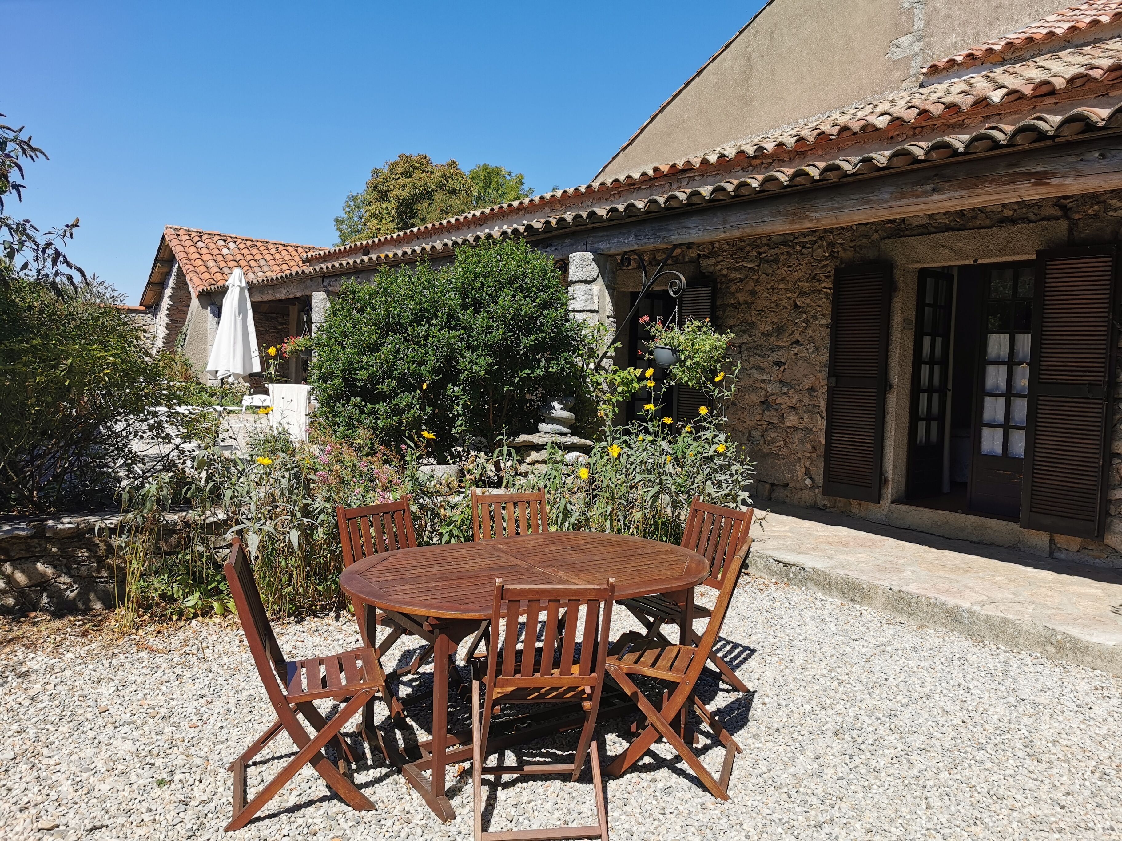 Un área de comedor al aire libre de estilo rústico, con una mesa y sillas de madera redondas, rodeada de grava y plantas.