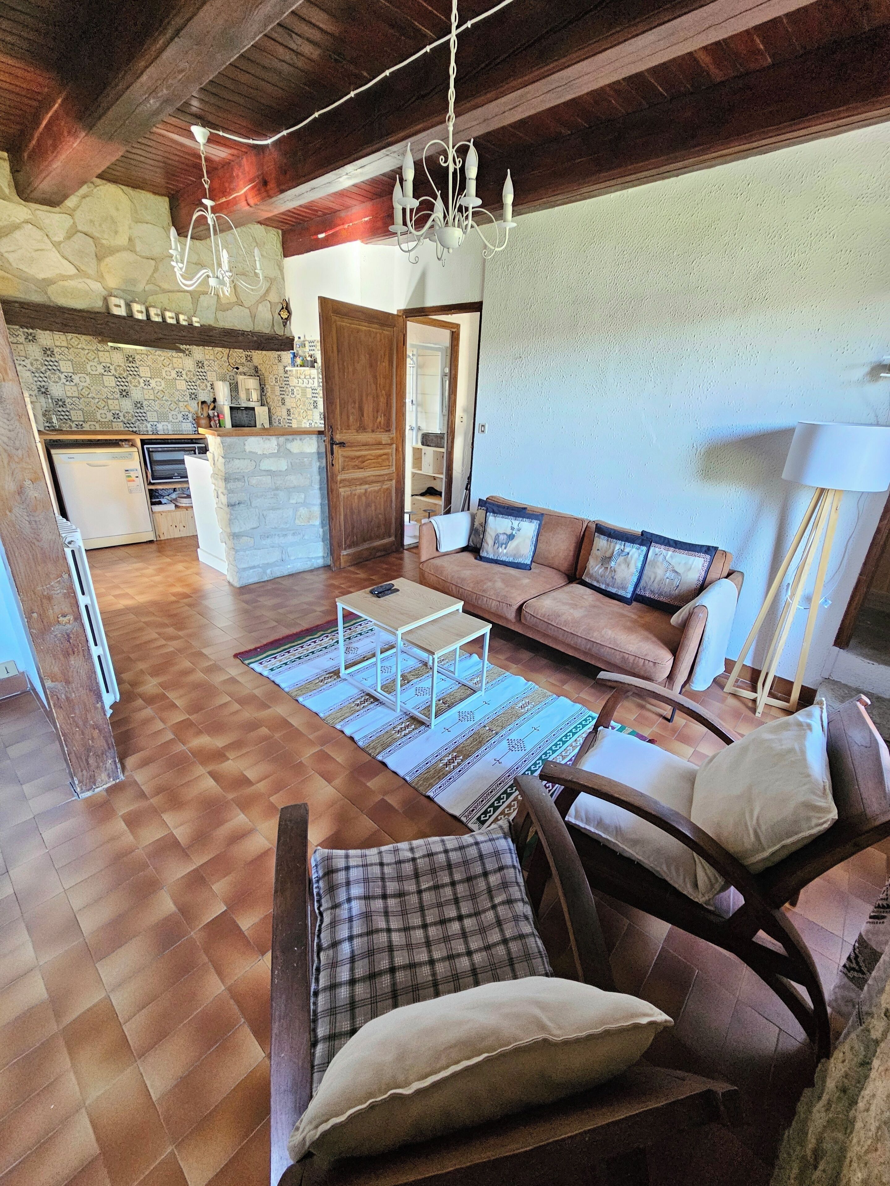 Un salon comprenant un canapé, des fauteuils, une table basse, un lustre, une cheminée et un coin cuisine avec un mur en pierre et des poutres en bois.