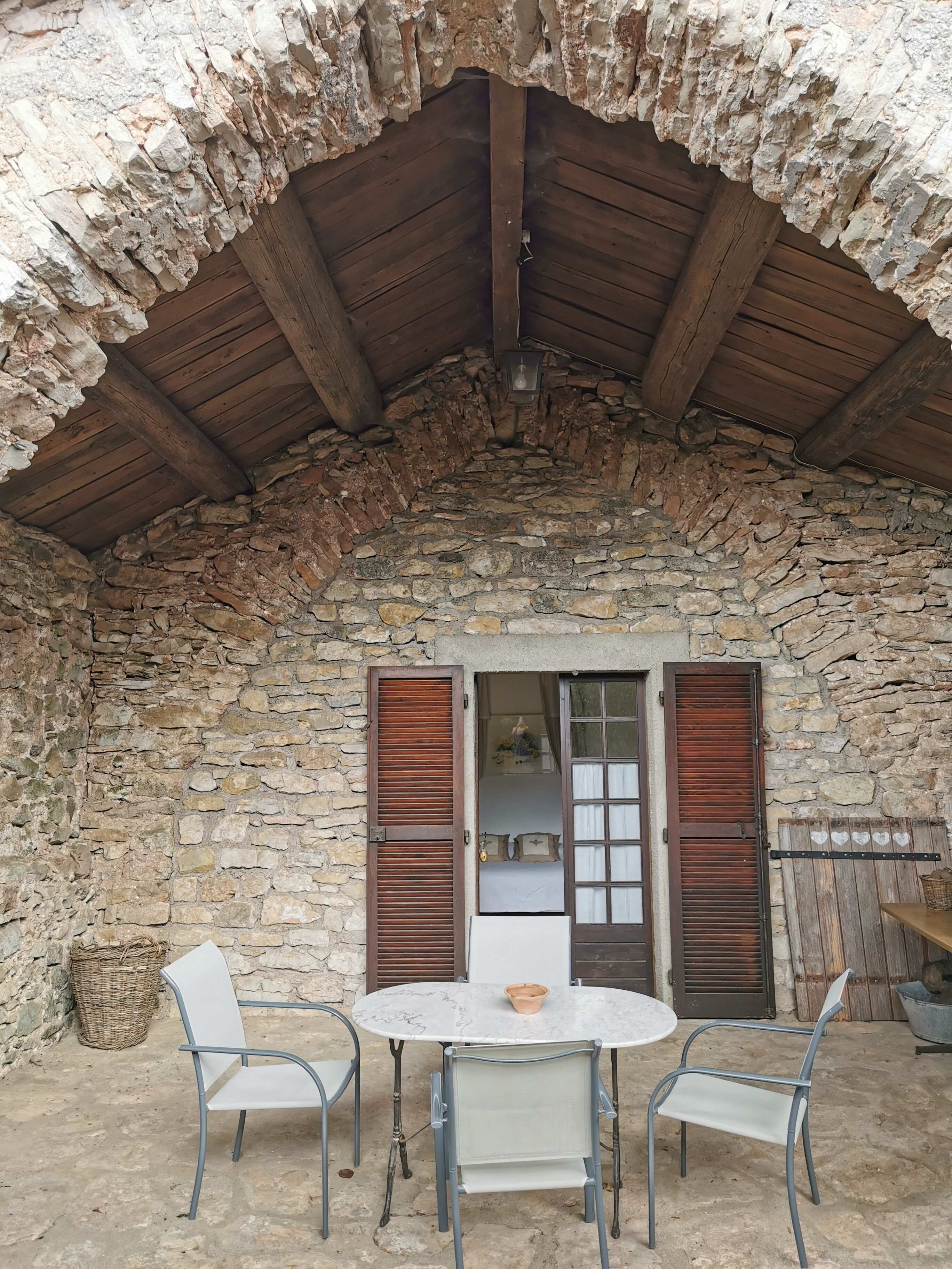 A stone-walled room with a wooden ceiling, a round table with chairs, and wooden shutters on the windows.