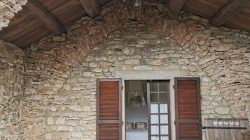 A stone-walled room with a wooden ceiling, a round table with chairs, and wooden shutters on the windows.