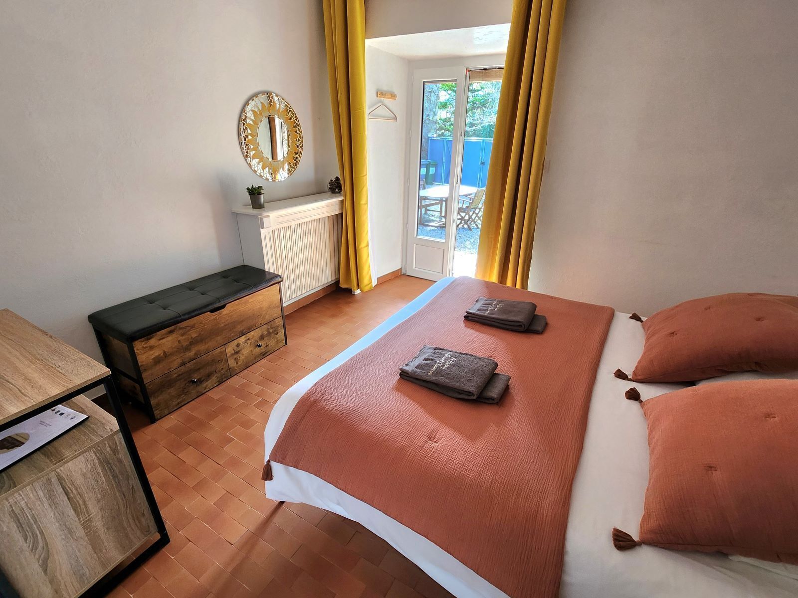 A bedroom with a bed, a chest of drawers, a bedside table, a mirror, and a view of the outside.