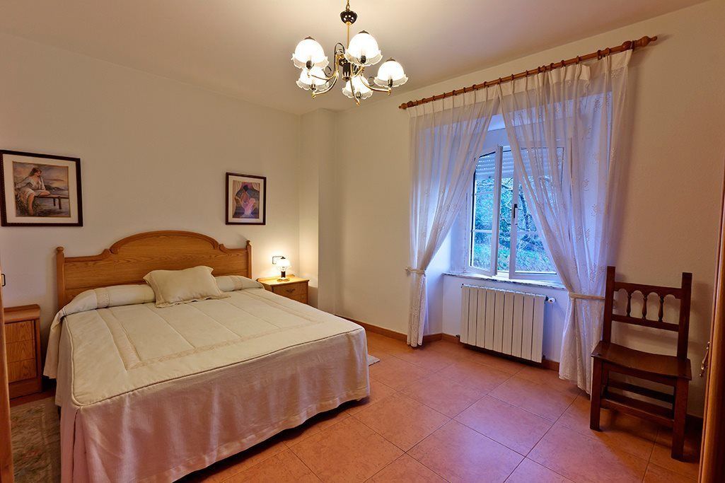 Un dormitorio con cama de madera, una mesita de noche, una silla, una ventana con cortinas translúcidas y un radiador.