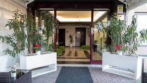 A hotel entrance with a checkered floor, two potted plants, a green chair, and a white bench. - Hotel Ayri (Medesano)