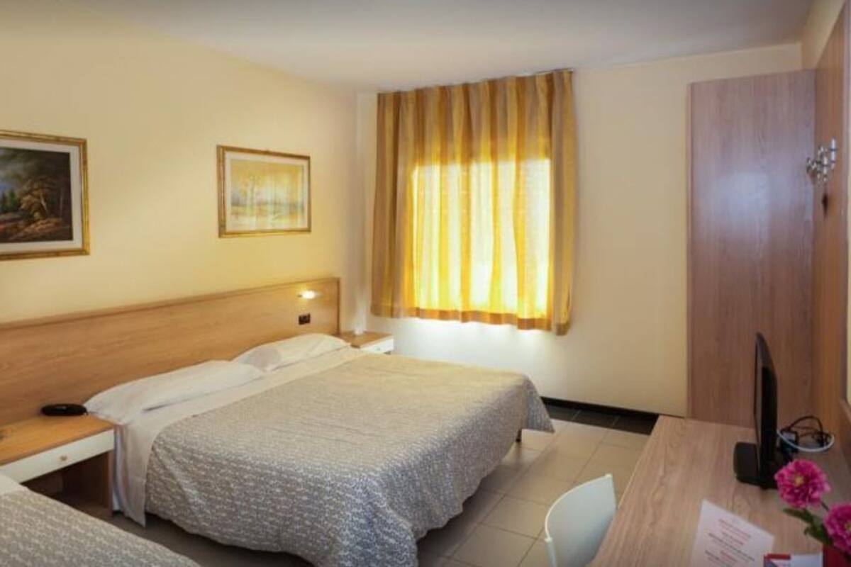A hotel room with a bed, a desk with a computer, a chair, a wardrobe, and a window with curtains.