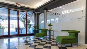 A hotel lobby with a checkered floor, two green armchairs, a potted plant, and a glass wall with the hotel name. - Hotel Ayri (Medesano)