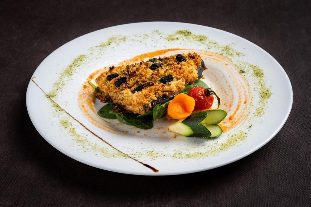 A white plate with a piece of food, possibly a fish fillet, topped with breadcrumbs and garnished with a sauce and vegetables.