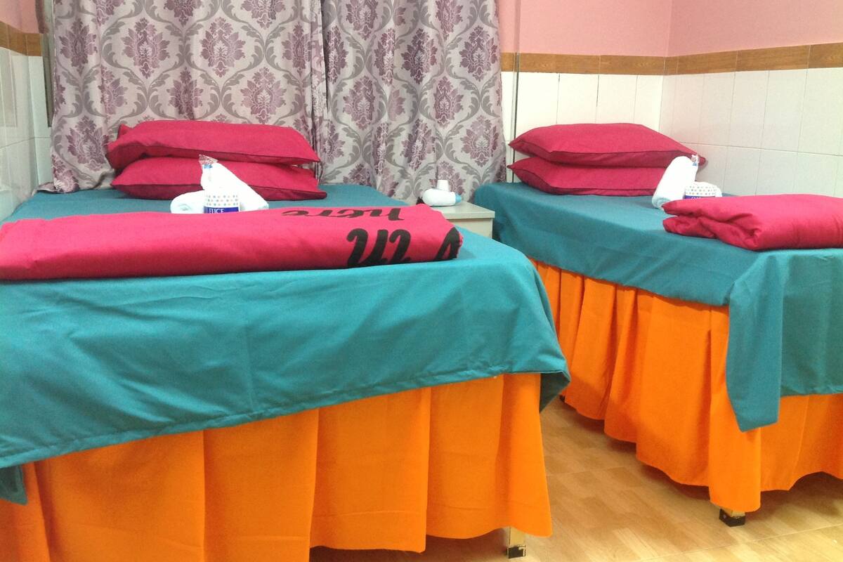 Two beds with teal and orange bedding, red pillows, and a patterned curtain in a room with wooden flooring.