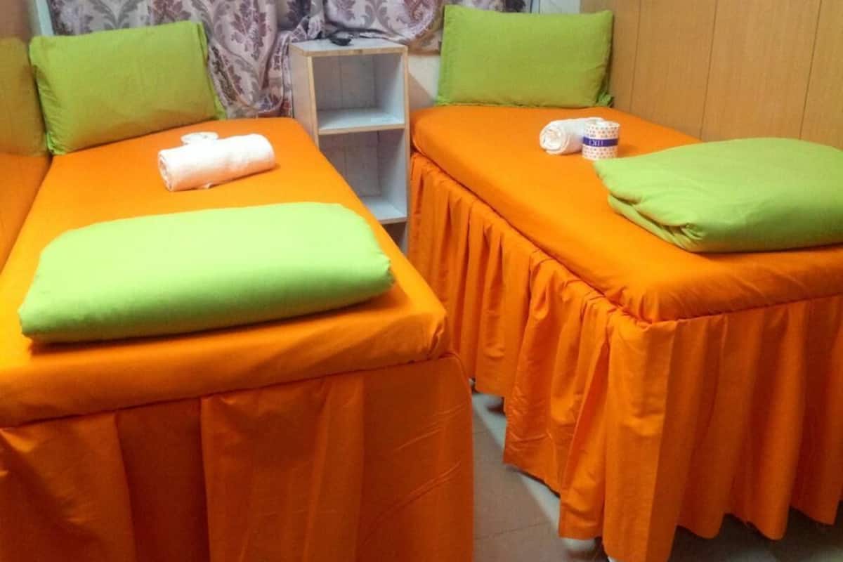 Two single beds with orange bedding and green pillows, a small shelf unit, and a patterned curtain in a room.
