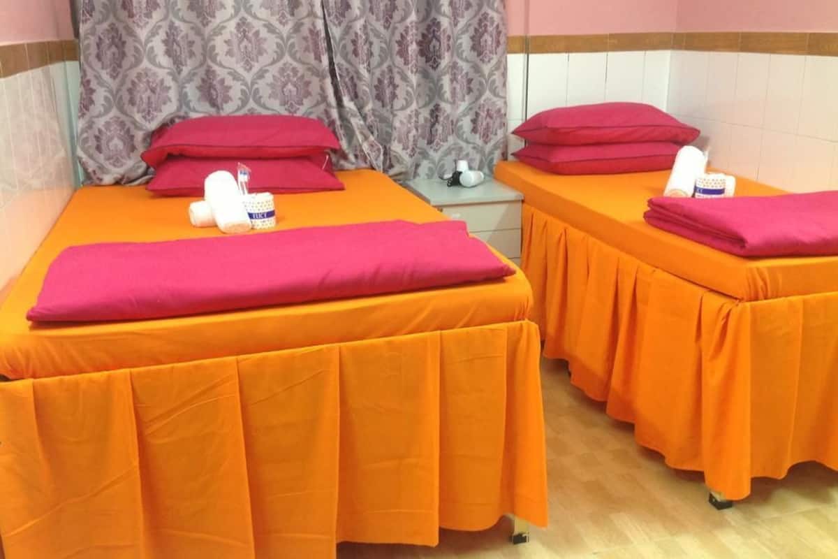 Two massage therapy beds with orange covers and red pillows, a patterned curtain, and a small bedside table with a bottle and a container.