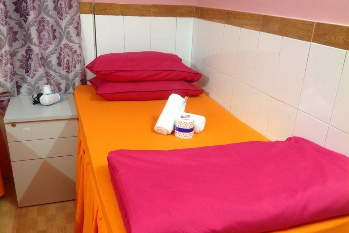 A single bed with an orange bedspread and two red pillows. A white bedside table with a lamp and a small item on it is visible.