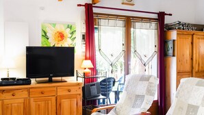 A living room with a TV, wooden cabinets, a painting, and a window with curtains. - Cozy Villa Studio with Mezzanine, Terrace, and Parking near Narbonne-Plage (Narbonne-Narbonne Plage-Narbonne Plage)
