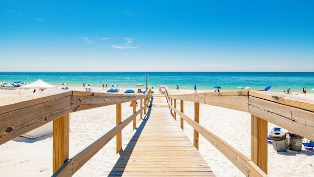 Een houten looppad dat naar een strand leidt, met parasols en mensen op het strand.