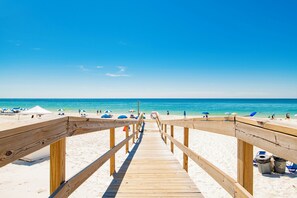 Un paseo marítimo de madera que lleva a una playa con sombrillas y gente.