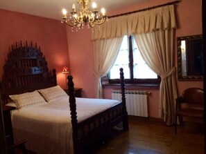 Un dormitorio con una cama con dosel, un candelabro, un radiador y una ventana con cortinas.