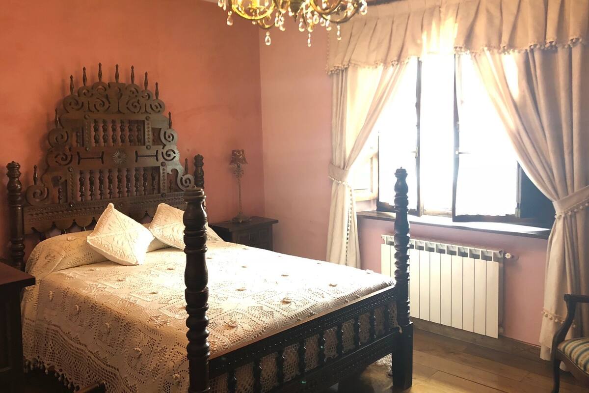 A bedroom with a four-poster bed, a chandelier, a radiator, and a window with curtains.