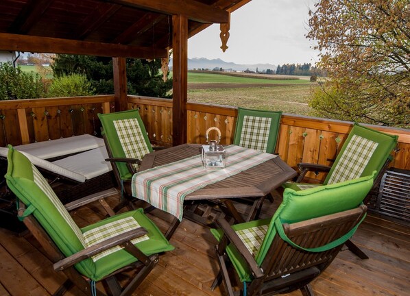 Une terrasse en bois avec une table et des chaises, d’où l’on peut admirer un champ et des montagnes en arrière-plan.
