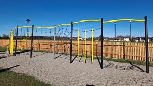 A playground with yellow and black play structures, including swings and climbing frames, set against a backdrop of a wooden fence and houses. - Ferienwohnung im 1.Stock mit überdachter Terrasse und See- und Bergblick (Chieming)