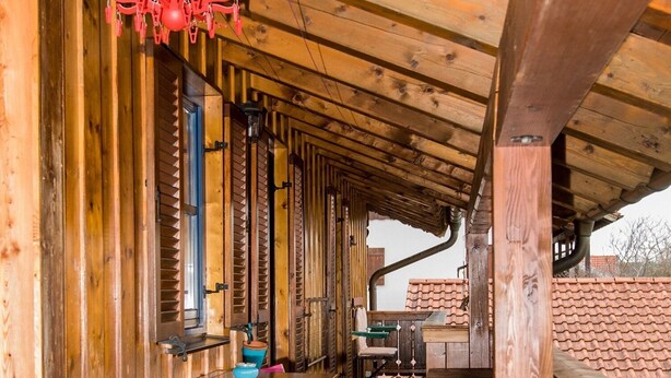 A wooden covered porch with a table and chairs, a hanging red decoration, and a view of a tiled roof in the background. - Ferienwohnung im 1.Stock mit überdachter Terrasse und See- und Bergblick (Chieming)