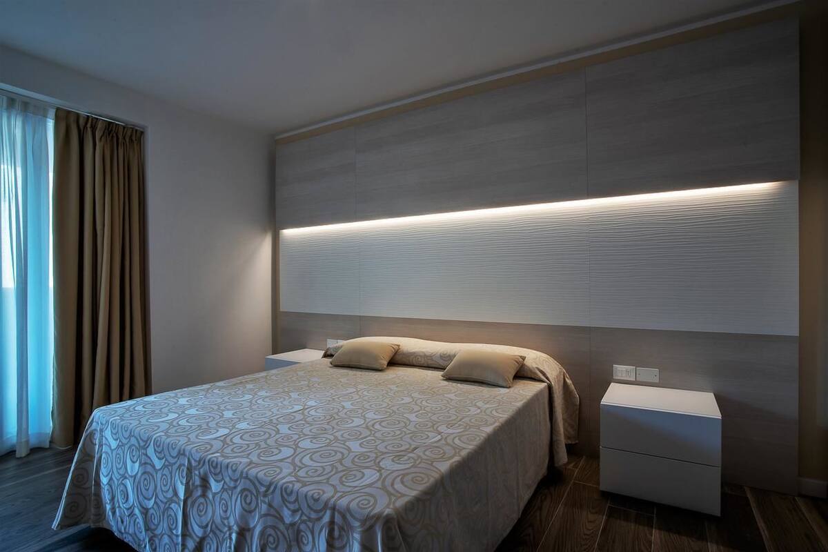 A modern hotel room with a large bed, a bedside table, and a headboard with integrated lighting.