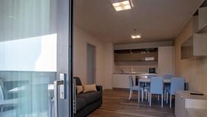 A modern apartment with a glass door leading to a balcony, a living area with a sofa, a dining area with chairs and a table, and a kitchenette with cabinets and appliances. - Aparthotel Miramare (Grado)